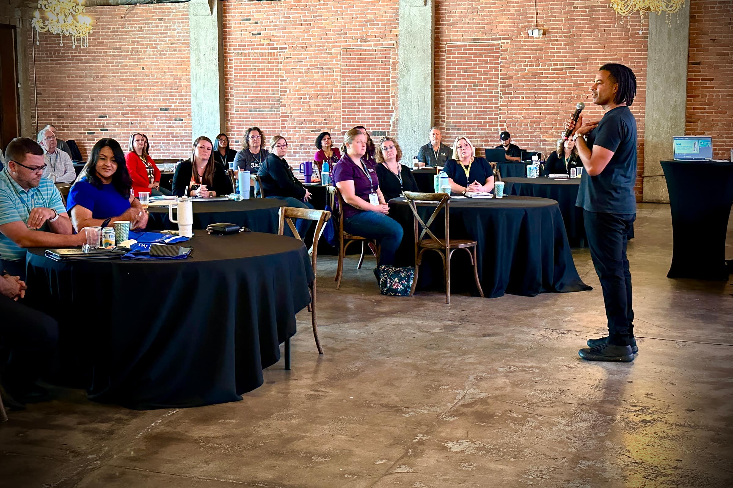 Antonio Neves speaking with a microphone to about twenty people seated at round tables in a brick walled room
